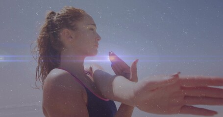 Stretching athlete pulling arm across chest at beach in dark top with pink trim, lens-flare streaks