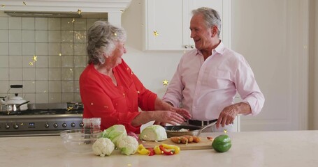 Chopping senior couple preparing meal in kitchen at island with vegetables, red blouse, pink shirt