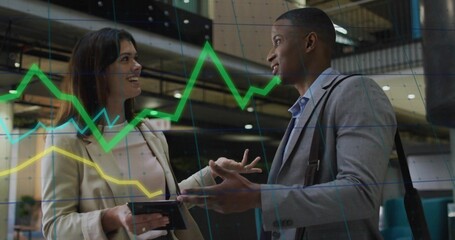 Discussing pair in business attire with tablet and shoulder bag under faint chart overlay in atrium