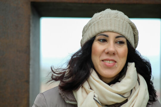 Close-up of a woman portrayed in a public park. The woman is thirty-nine years old, has long black hair and wears a beige coat, scarf and hat