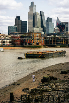 A mudlarker on the South Bank walks past a barge on the river Thames as the skyscrapers of the City of London stand in the distance
