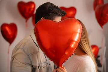 A man and woman playfully posing with a red heart balloon in a room decorated for Valentine's Day. Romantic surprise, love and celebration concept.