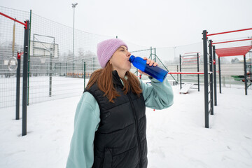 Girl drinks from a bottle on a snowy sports ground in winter
