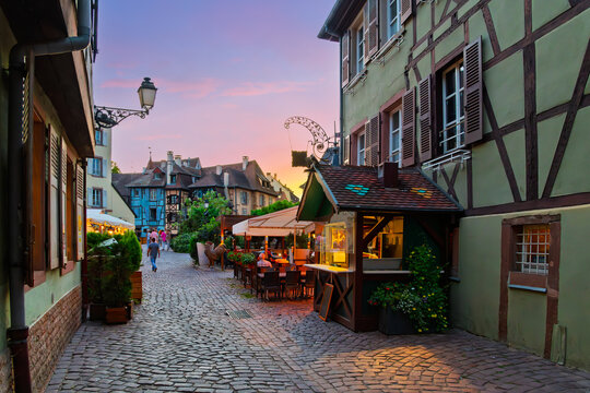 Cobblestone streets of sidewalk cafes and shops illuminated at twilight in the Petite Venise district of the Alsatian city of Colmar, France.