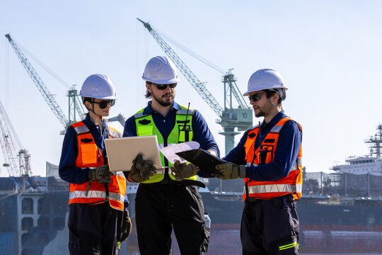 group of industrial engineers workers in a refinery - oil and gas processing equipment and machinery, engineers collaborate with a laptop, blueprint, and digital tablet at the oil storage tanks site. - Powered by Adobe