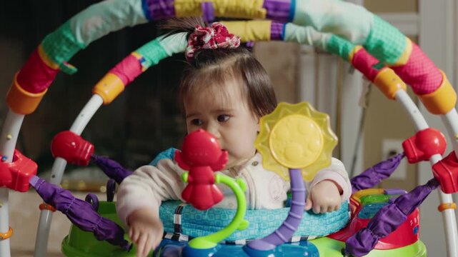 Baby girl bouncing playfully in a colorful activity jumper, making a funny face while exploring the bright toys around her