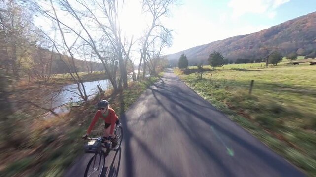 A mature woman rides her bike on a road next to a river in West Virginia as autumn colors fill the landscape and the sun shines brightly.
