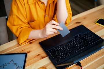 Woman holding credit card near laptop with stock market charts on wooden table. Focused financial activity supporting wealth management.