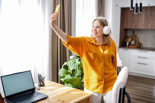 Smiling business woman taking selfie with smartphone in home office wearing headphones. Freelance finance work and personal brand communication.