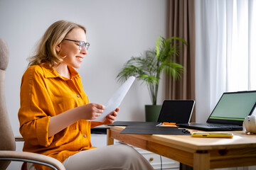 Smiling woman sitting at desk and holding document in bright room. Happy professional successful with project results or job offer.