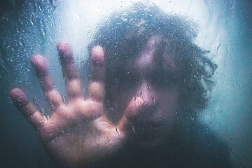 A person presses against a fogged glass surface, creating a sense of isolation and introspection, with water droplets adding to the mood.