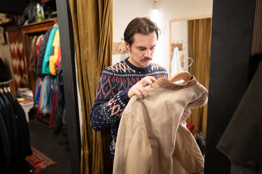 Fashionable mustachioed man picking fancy outerwear in second hand clothing shop. Hipster male in apparel boutique holding vintage jacket on hanger and examinees item quality, mindful consumption.