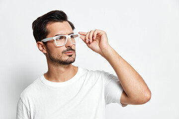 Thoughtful young man wearing white glasses adjusts frames while looking to the side, dressed in a...