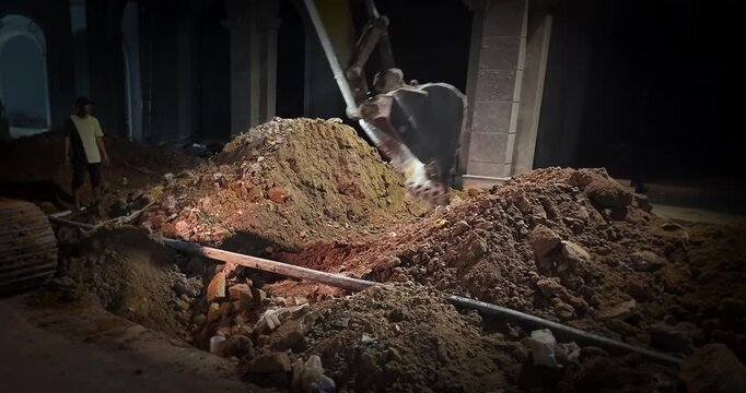 Excavator digging into a large pile of dirt at a construction site in front of a building at night.