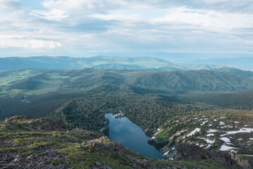 Top view from rocky precipice edge to blue lake in woody hilly terrain against mountain range silhouette far away. Cloudy sky reflected in smooth water surface of long forest lake among green hills.