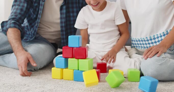 Family parents and daughter play blocks together. On a carpeted floor, a bright pyramid of plastic cubes; hands steady pieces, building focus and motor skills. Playful family bonding at home.