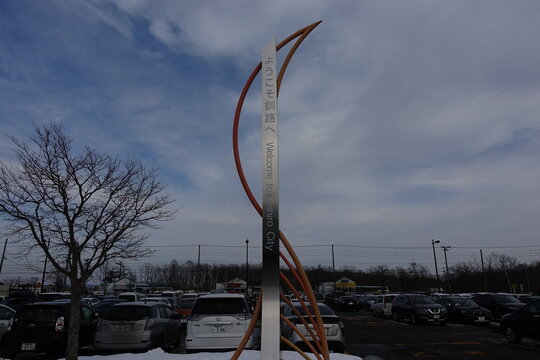 "Welcome to Kushiro City" metal monument sign in parking lot with snow and cars.