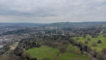 Fototapeta premium High aerial view over Bath showing a large park with intersecting paths, scattered trees, surrounding neighbourhoods of historic houses, and rolling green hills under an overcast sky.