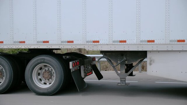 POV Close tracking shot of semi-truck trailer and rear wheels moving on the road, showing suspension, landing gear, and transport details in motion