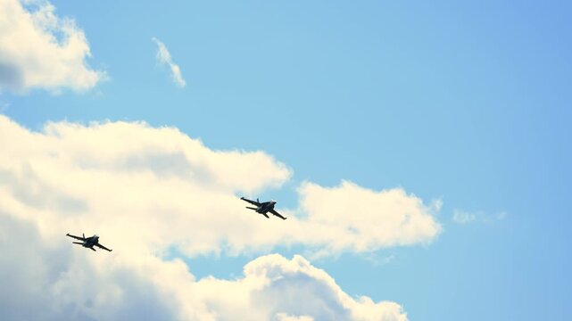 Two military fighter jets fly through a bright cloudy sky in loose formation, leaving subtle trails as they move together across open airspace. POV