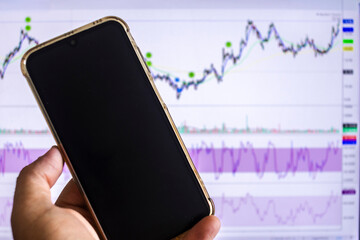 Man's hand holding a smartphone in front of a screen with a stock market chart, symbolizing mobile trading, investment, and financial analysis in the digital age.