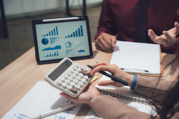 Financial Analysis Collaboration: Two professionals collaborate, examining intricate charts and figures on a tablet, while one carefully operates a calculator.