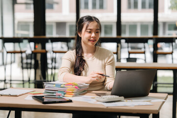 Fototapeta premium Focused in Office: A focused professional, engages in her work at her desk, utilizing a laptop and organizing documents amidst the modern office ambiance.
