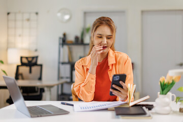 Obraz premium Asian woman wear orange shirt use laptop pc computer hold mobile cell phone sit alone at table in coffee shop cafe restaurant indoors work or study Freelance office business concept 