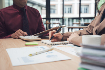 Analyzing Data: A focused perspective of two business people working in the office, meticulously examining data reports, utilizing a calculator, and capturing thoughts in a notebook.