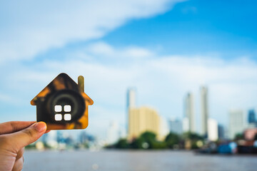 Hand holding metallic house model with city skyline background and river view, representing urban...