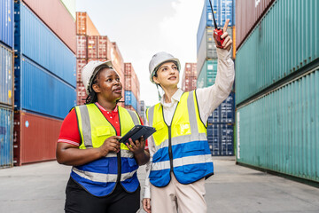 Two diverse female logistics workers in safety vests and hard hats using digital tablet and radio at shipping port terminal. Professional team discussing cargo operations in container yard background