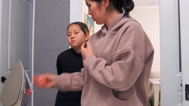 Mom ironing clothes on a board while explaining safety rules to her daughter. Parent shows careful handling of hot iron and teaches household responsibilities.