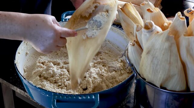 Woman's hands spreading masa dough on a corn husk to make traditional tamales in a rustic kitchen