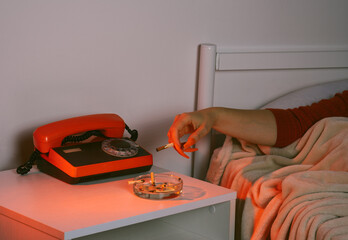 Hand with false nails holding cigarette near retro phone on bedside table at night