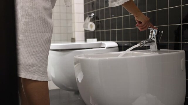 A woman in a white coat turns on the water bidet with a chrome faucet in a tiled bathroom. The wall has a mix of dark and light tiles, creating a stylish contrast.
