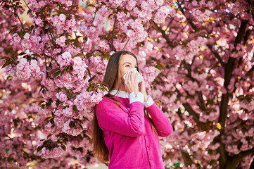 Woman allergic suffering from seasonal allergy at spring in blossoming garden at springtime. Woman sneezing and blowing nose using nasal handkerchief in front of blooming tree. Spring allergy concept.