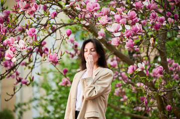 Woman allergic using medical nasal drops, suffering from seasonal allergy at spring in blossoming garden. Young woman treating runny nose in front of blooming tree outdoors. Spring allergy concept.