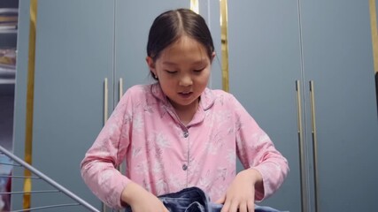 Young Asian girl in pink pajamas organizing and hanging laundry on an indoor drying rack. Household chores, daily routine, self-care skills, tidiness, and family responsibility.