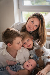 Obraz premium A mother embraces her three young sons, smiling warmly by the window as they sit together on the bed
