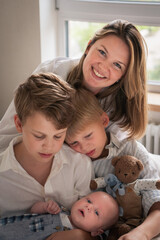 Obraz premium A mother embraces her three young sons, smiling warmly by the window as they sit together on the bed