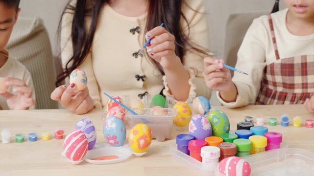 Easter egg painting. A mother and kids celebrate Easter indoors, using a paintbrush to decorate eggs near a festive basket.