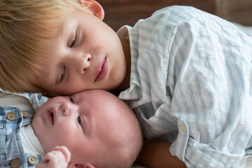 Older brother lies close to newborn baby on bed, neutral colors, gentle facial expressions, calm...