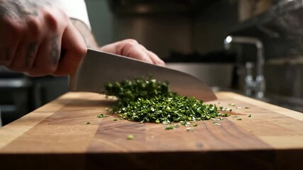 A chef's hands meticulously chopping fresh green herbs with a sharp knife on a wooden cutting board in a professional kitchen setting.