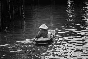 Obraz premium A lone rower wearing a traditional conical hat glides peacefully through the calm canal waters. The black and white tones capture a timeless moment of local life along the river.