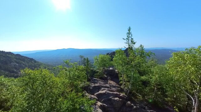 panoramic view of the rugged mountain peaks and dense green forests of the Southern Urals in Russia on a bright, sunny summer day. The clear blue sky and natural rock formations provide a perfect back