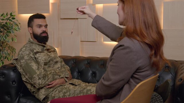 Middle aged female psychologist hypnotizing African American male soldier in military uniform using pendulum, to create state of focused attention during treatment of psychological disorder.