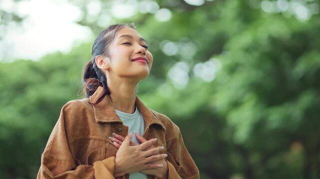 Peaceful young Asian woman with eyes closed and hands on her heart, expressing gratitude, relief, or mindfulness in a green park environment, Girl connects with her emotions during a reflective moment