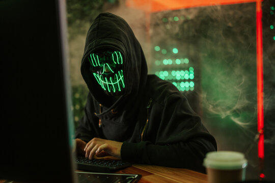 Hacker wearing neon mask typing on computer in dark room - Powered by Adobe