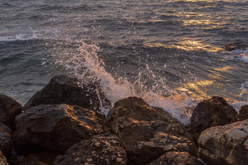 Mediterranean sunset scene with sea waves splashing against coastal rocks near Athens, Greece. Soft golden reflections on water and layered clouds create a peaceful natural background.