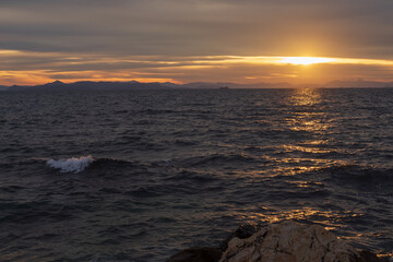 Moody sunset scene with golden light reflecting on dark Aegean Sea waves under layered clouds. Peaceful seascape near Athens, Greece, suitable for travel, nature and background use.